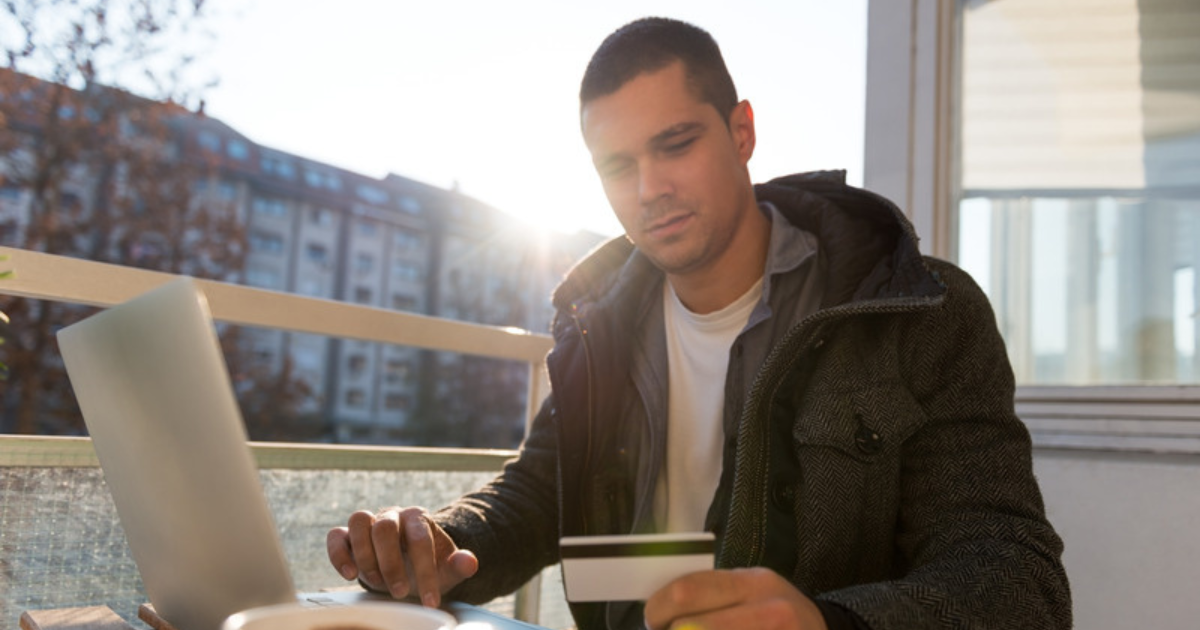 Image of young, gen z, male adult sitting at a table in front of a laptop with a card in his hand, considering his finances